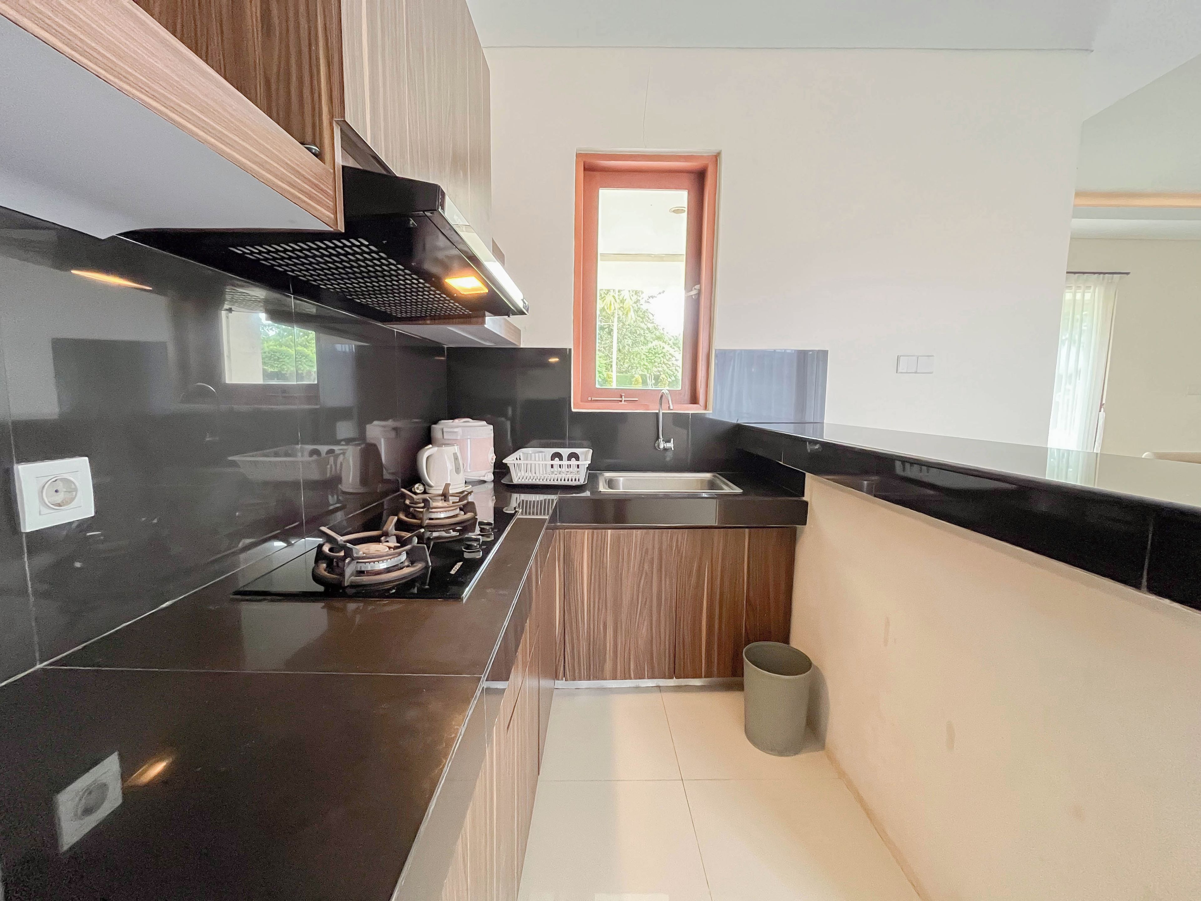 A modern kitchen with black countertops, wooden cabinets, a stainless steel range hood, a window, and a small trash can.