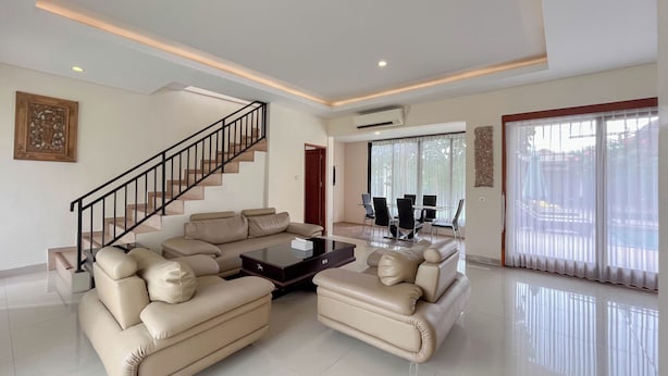 A spacious living room with a beige sectional sofa, a coffee table, a staircase, and a dining area with a glass table and chairs. - Spacious And Homey 3Br At Villa Casa De Uli (Jimbaran)