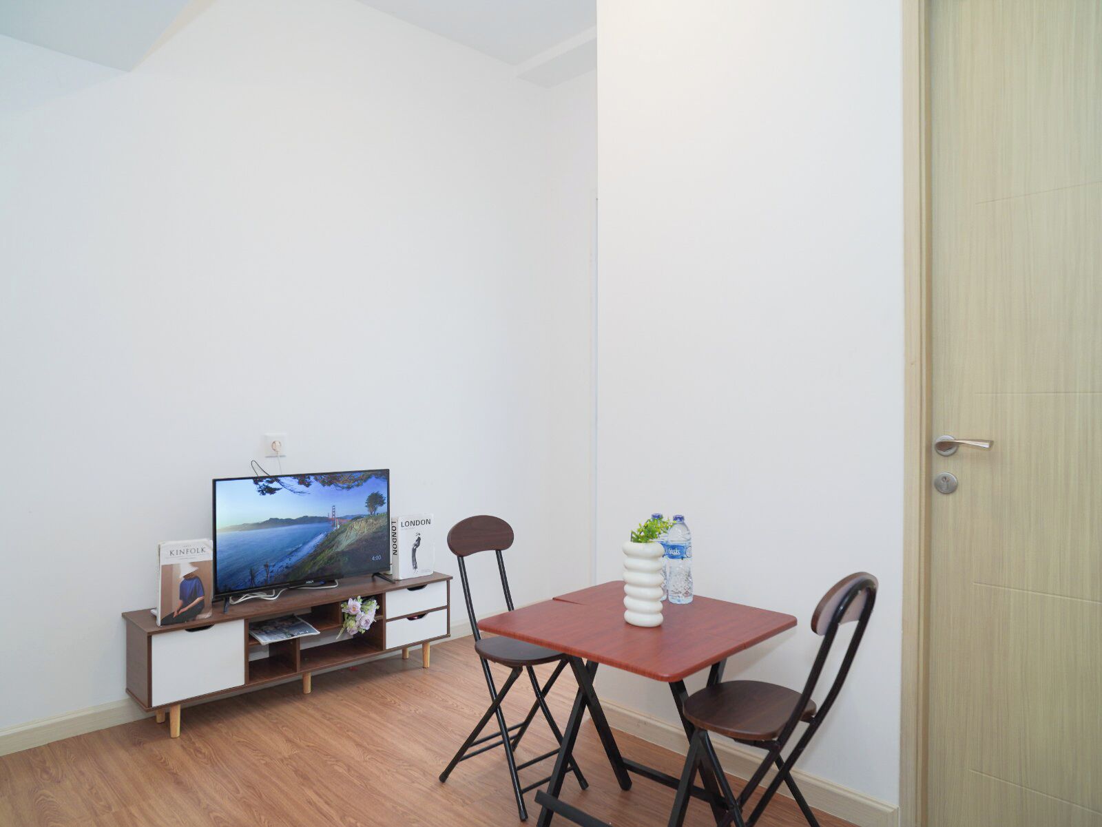 Una stanza piccola con pavimento in legno, un televisore su un supporto in legno, un tavolo con sedie e una porta.