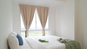 A bedroom with a bed, two windows with curtains, a view of greenery outside, and a green plant on the bed.