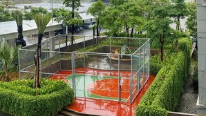 A basketball court with a red surface, surrounded by a metal fence and lush greenery.