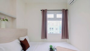 Ett hotellrum med en säng, ett fönster med gardiner, en liten krukväxt och en väggmonterad luftkonditioneringsanläggning.