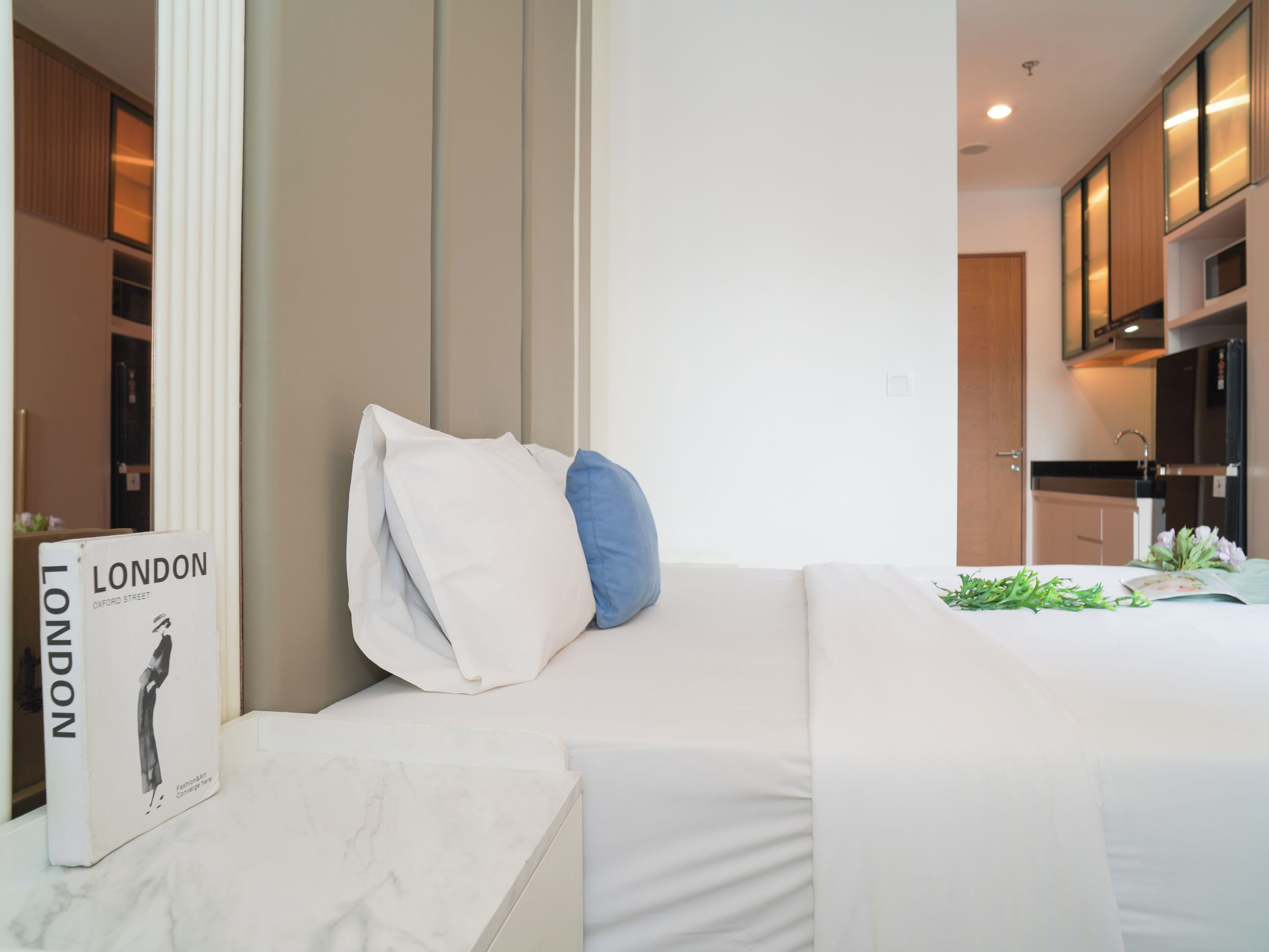 A modern hotel room with a neatly made bed, a small table with a book, and a kitchen area in the background.