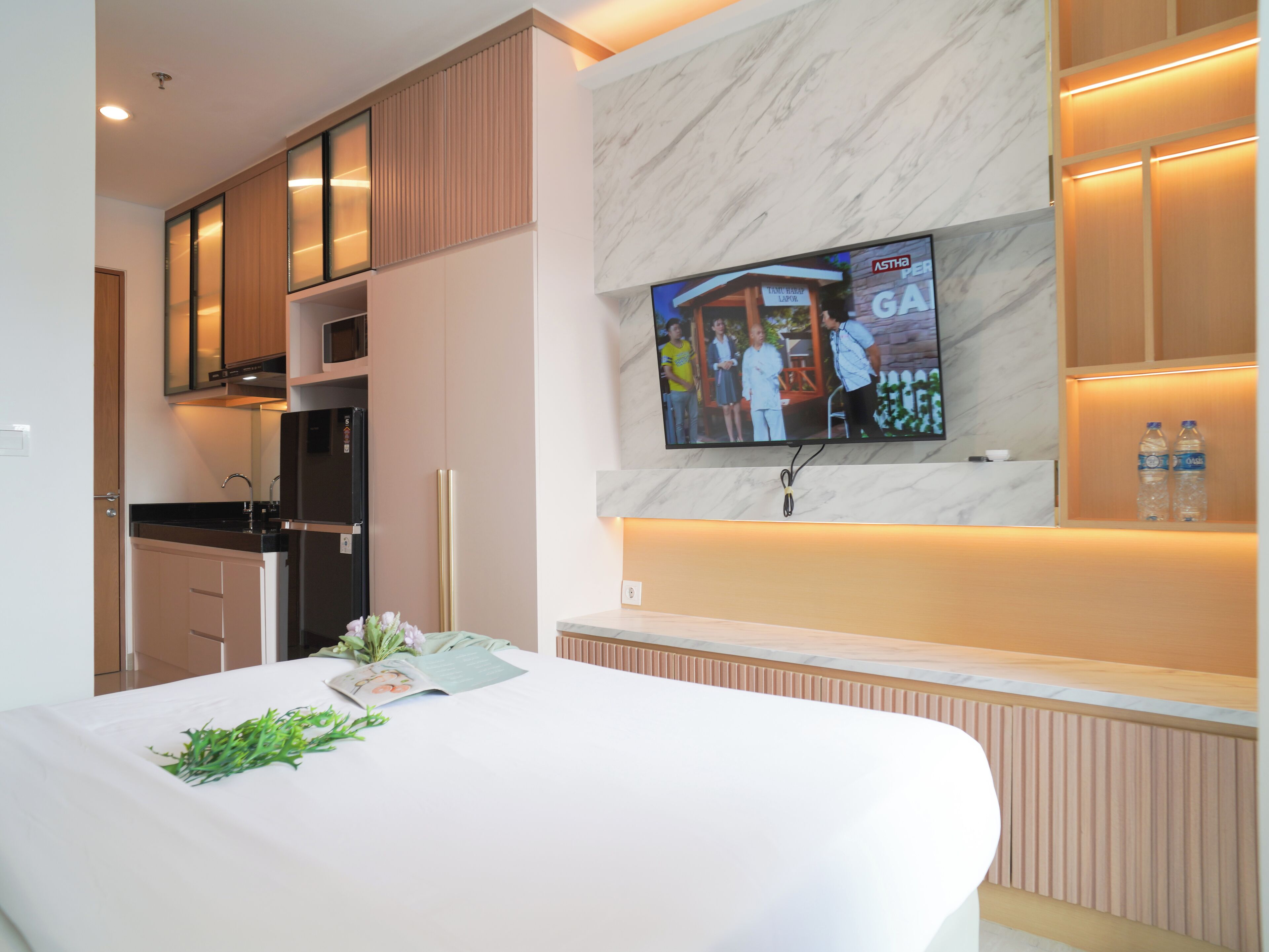 Ein modernes Hotelzimmer mit einem großen Bett, einem an der Wand montierten Fernseher, einer Küchenzeile und einer Minibar.