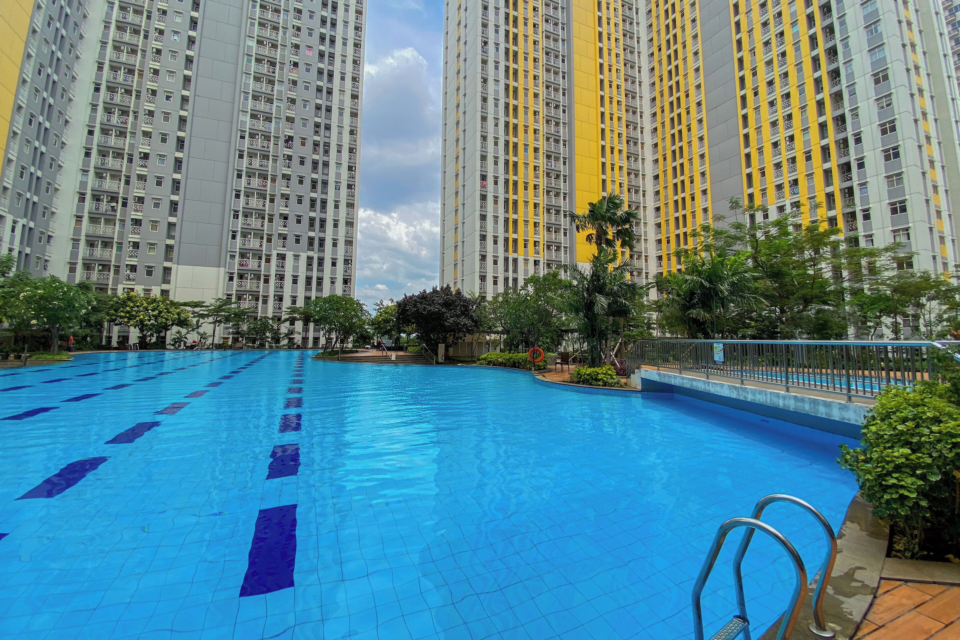 A swimming pool with clear blue tiles and lane dividers, surrounded by greenery and tall residential buildings in the background.
