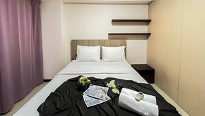 Una habitación de hotel con cama, mesita de noche, ventana con cortinas y estante fijado a la pared.