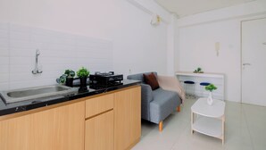 A modern kitchen with a sink, stove, and cabinets, a grey sofa, a small white table, and a white door. - Compact With Minimalist Designed 2Br At Bassura City Apartment (Jakarta)