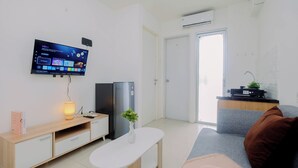 A modern living room with a flat-screen TV mounted on the wall, a wooden TV stand, a white table, a grey sofa, and a brown cushion. - Compact With Minimalist Designed 2Br At Bassura City Apartment (Jakarta)