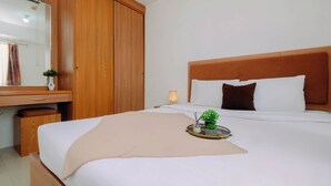 A neatly arranged hotel room with a wooden wardrobe, a bedside table with a lamp, a bed with white linens, and a small plant on a tray. - Serene And Warm 2Br At Apartment Bassura City (Jakarta)