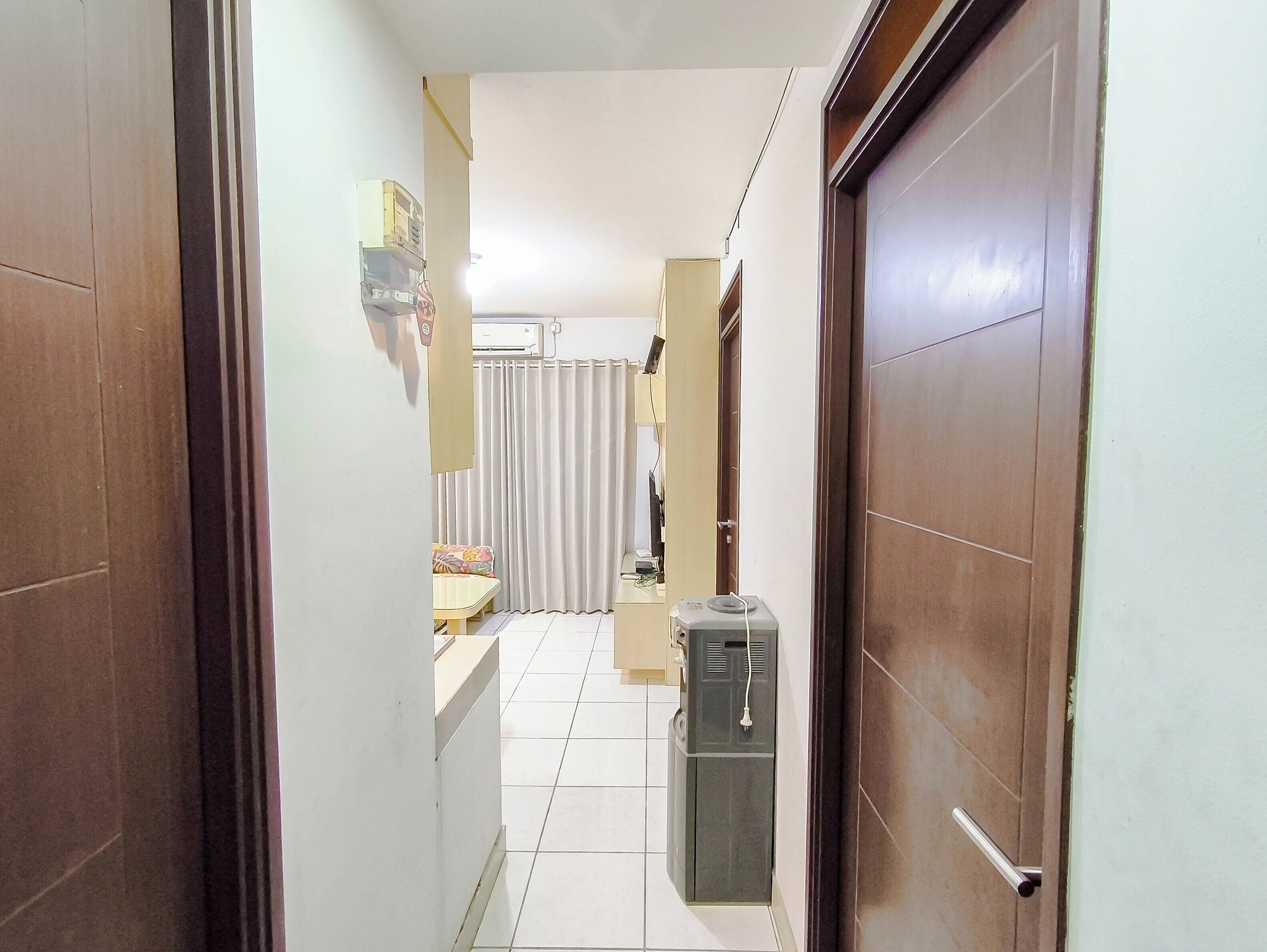 A narrow hallway with a tiled floor, a refrigerator, a small table with a floral cushion, and a door with a handle.