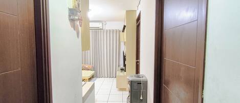 A narrow hallway with a tiled floor, a refrigerator, a small table with a floral cushion, and a door with a handle.
