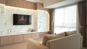 A modern living room with a flat-screen TV mounted on a marble wall, a sofa, and a large window with curtains. - Calm Tone 1Br At Tokyo Riverside Pik 2 Apartment (Teluknaga)