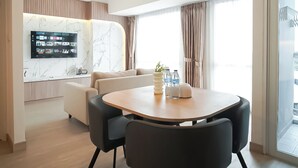 A modern hotel room with a marble accent wall, a flat-screen TV, a sofa, a dining table, and a balcony with a view. - Calm Tone 1Br At Tokyo Riverside Pik 2 Apartment (Teluknaga)