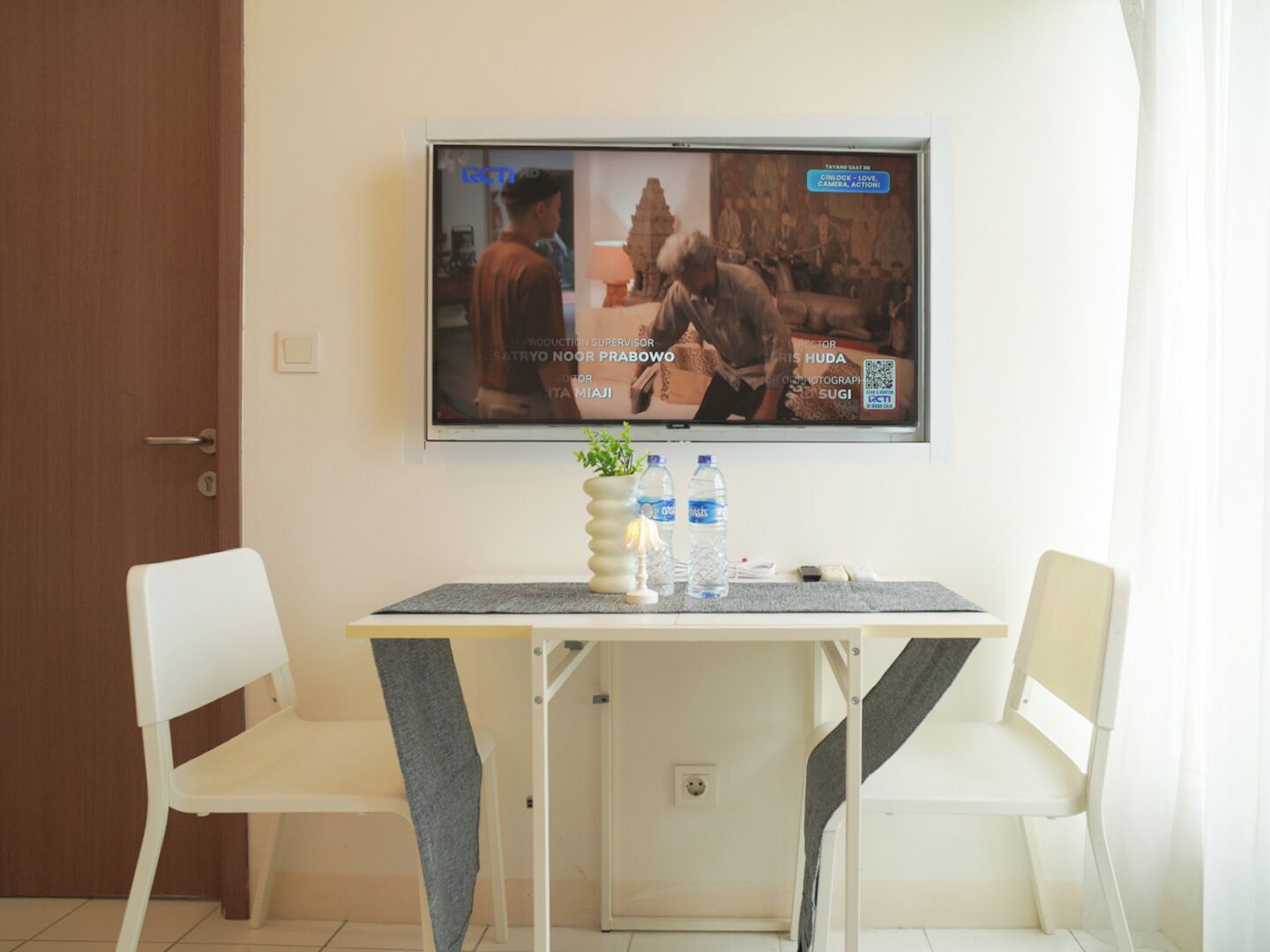 A room with a television mounted on the wall displaying a movie scene, a small table with two chairs, and a potted plant.