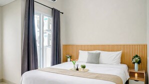 A hotel room with a large bed, wooden headboard, bedside tables with lamps, and a window with curtains. - Charming 1Br Apartment Lrt City Sentul - Royal Sentul Park (Bogor)