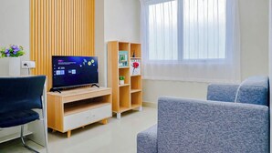 A modern living room with a flat-screen TV, wooden shelving, a comfortable sofa, and a large window with sheer curtains. - Charming 1Br Apartment Lrt City Sentul - Royal Sentul Park (Bogor)