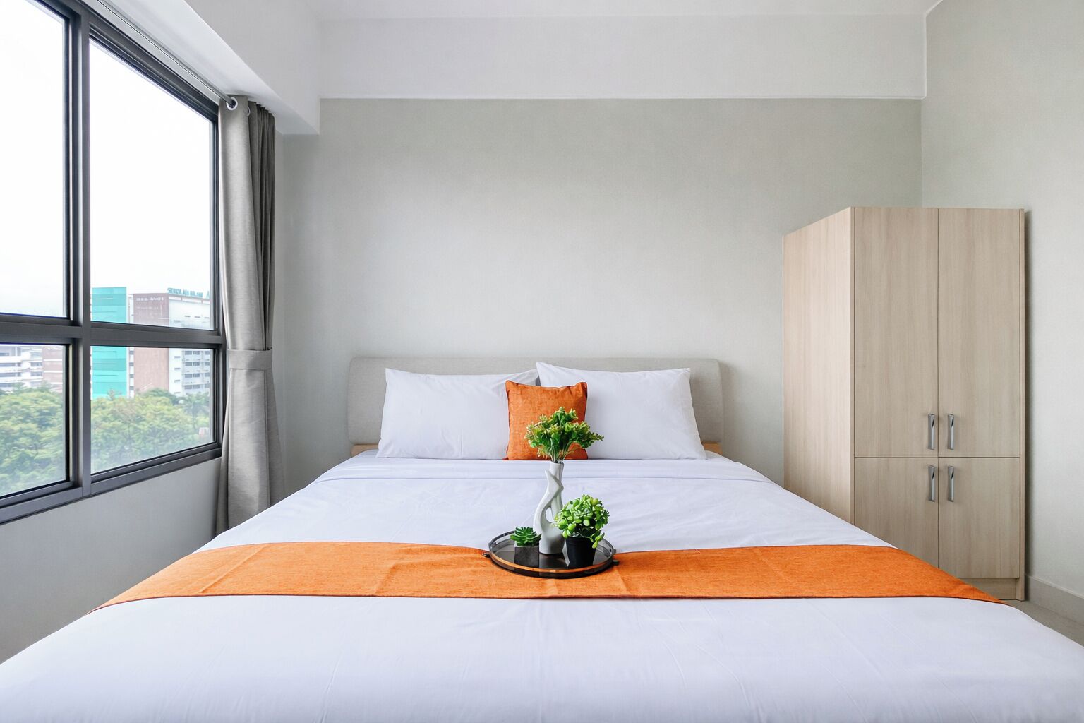 A neatly made bed with white bedding, an orange throw blanket, and a small potted plant on a bedside table.