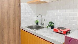 A kitchen with a white tiled backsplash, wooden cabinets, a stainless steel sink, a red portable stove, and two potted plants. - Modern Studio Apartment Near Sicc At Royal Sentul Park (Bogor)