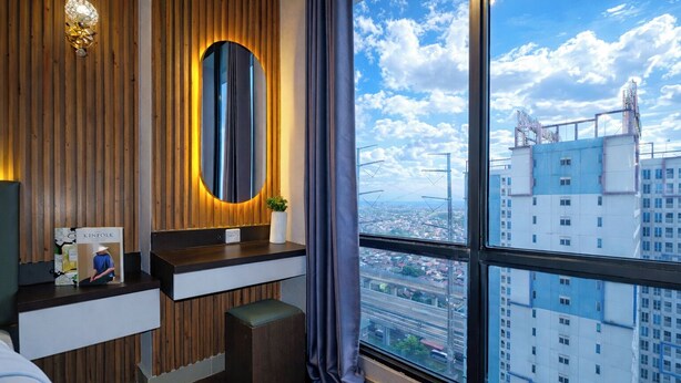 Een moderne hotelkamer met een houten wand, een groot raam met uitzicht op de stad, een kaptafel met spiegel en een klein bankje.