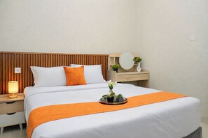 A neatly made bed with white bedding and an orange throw, a bedside table with a lamp, a small mirror, and a nightstand with a power outlet. - Tranquil 1Br At Royal Sentul Park Apartment (Bogor)