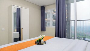 A hotel room with a large bed, a small table with a vase of flowers, a window with curtains, and a view of a building and greenery outside. - Tranquil 1Br At Royal Sentul Park Apartment (Bogor)