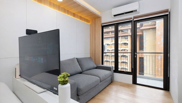 A modern living room with a grey sofa, a flat-screen TV, a white vase with a plant, and a large window with a view of a building. - Tranquil Studio At Tamansari Semanggi Apartment (Jakarta)