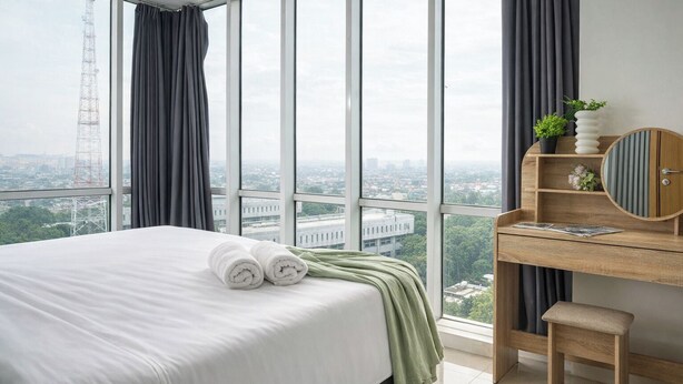 A hotel room with a large bed, a wooden desk, a small stool, and a view of the city through large windows. - 2Br Apartment With Service Room And Sofa Bed At Woodland Park Residence (Jakarta)
