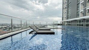 Una piscina infinita en la azotea con dos sillas de descanso, vistas a la ciudad y un edificio moderno al fondo.