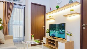 A modern living room with a sofa, a coffee table, a TV, and a wooden cabinet with shelves. - Comfortable And Contemporary 2Br At Apartment Transpark Cibubur (Bekasi)