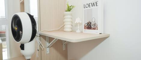Un taburete de madera, un escritorio blanco con una lámpara blanca, un ventilador blanco y un cuadro en la pared.