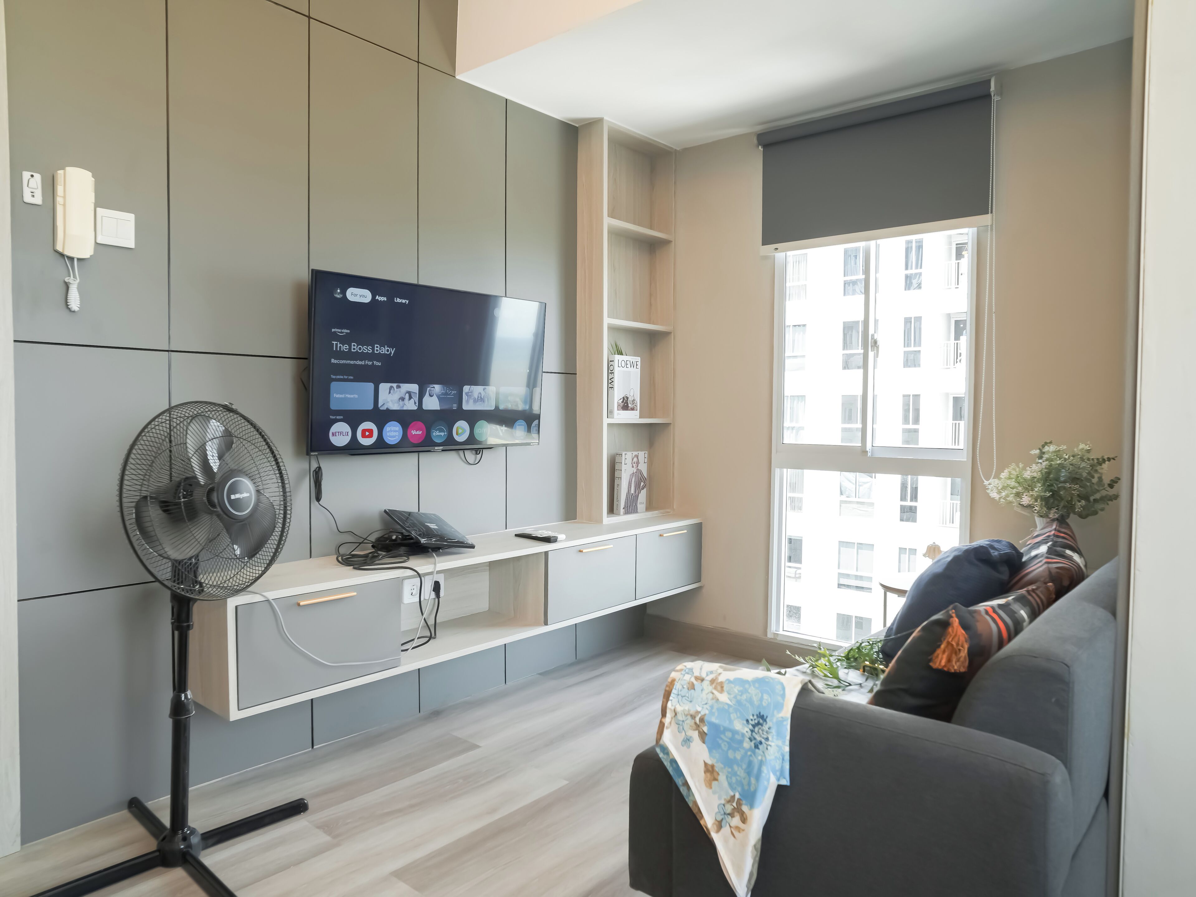 Ein modernes Wohnzimmer mit Flachbildfernseher, Standventilator, Sofa und Fenster mit Blick auf Gebäude.