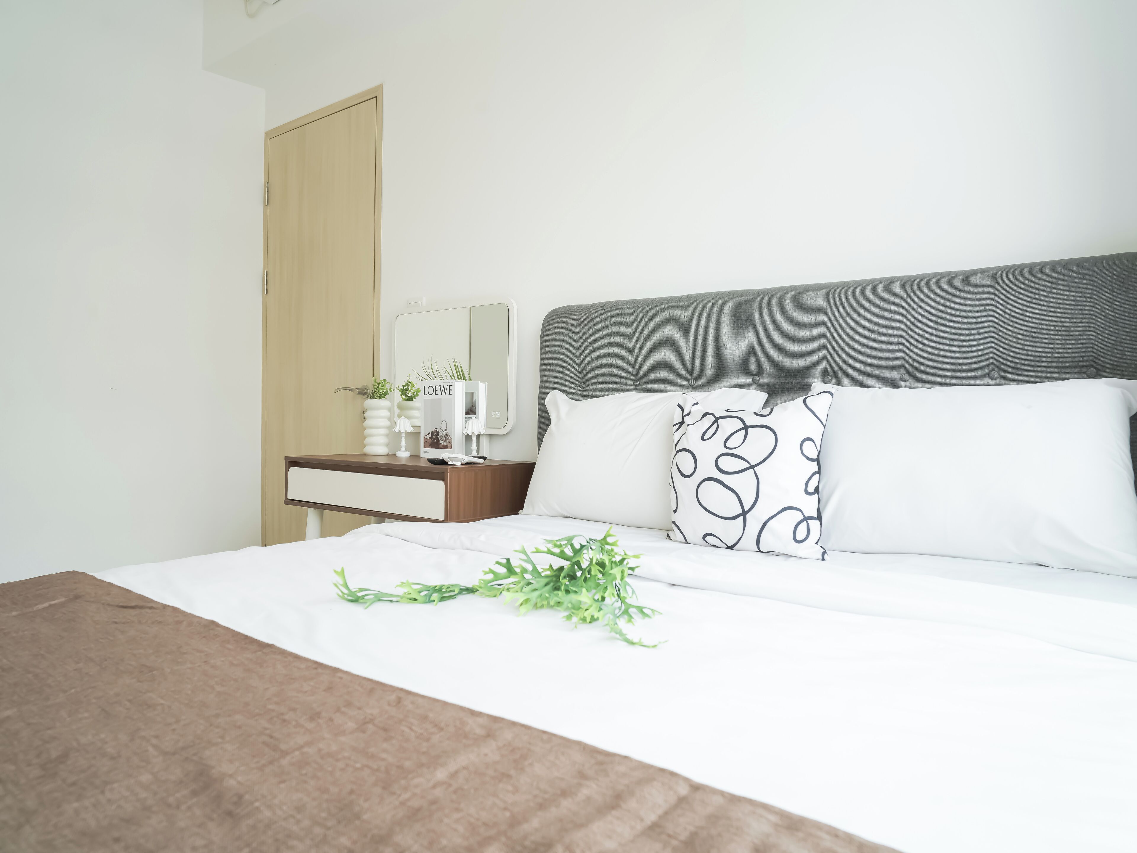 A neatly made bed with white and black patterned pillows, a bedside table with a mirror, and a small potted plant.