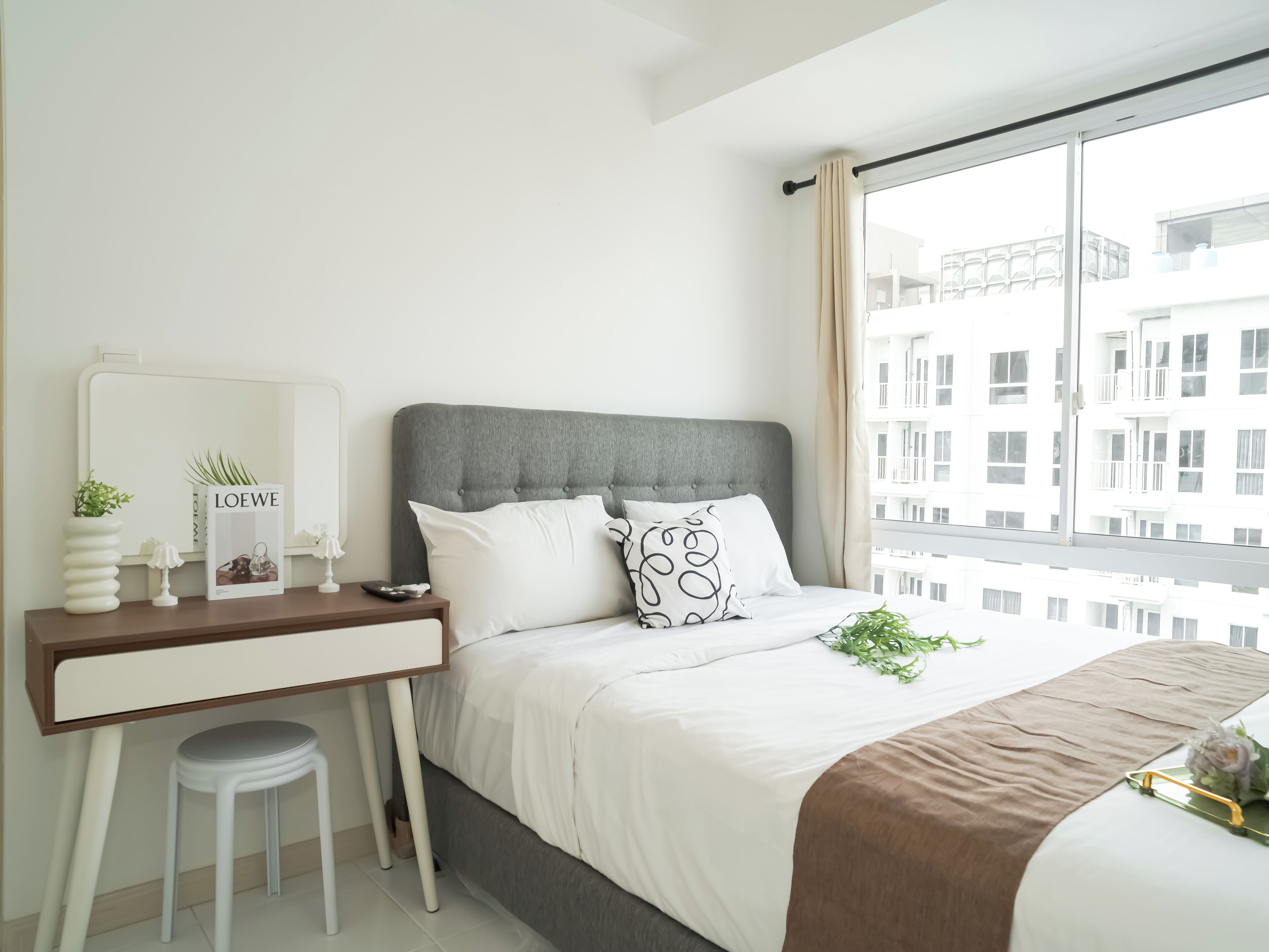 Un dormitorio moderno con cabecera tapizada en gris, ropa de cama blanca, un pequeño escritorio con espejo y vistas a edificios a través de amplios ventanales.