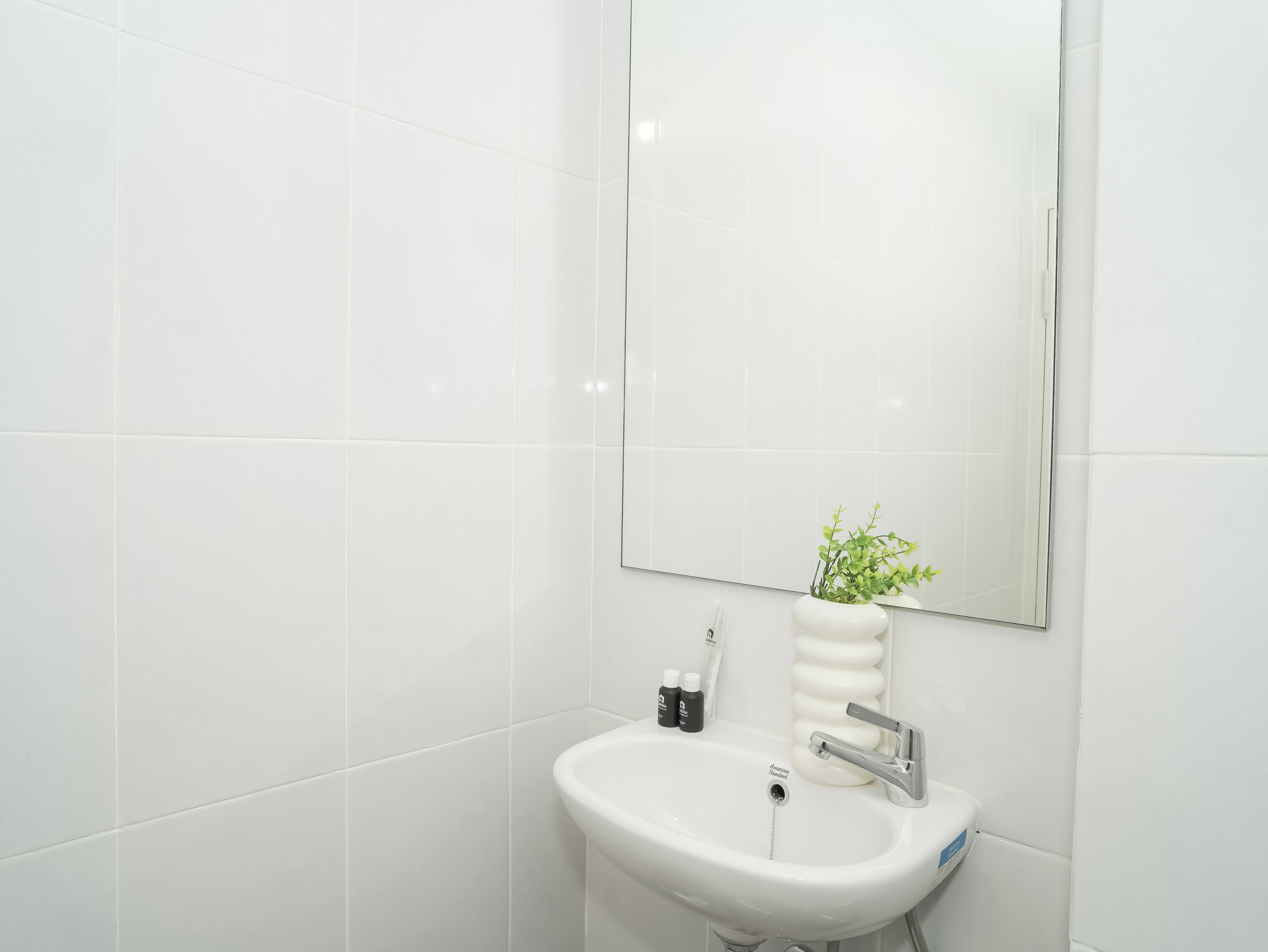 Un baño con baldosas blancas, un lavamanos blanco, un espejo y una pequeña planta en maceta.