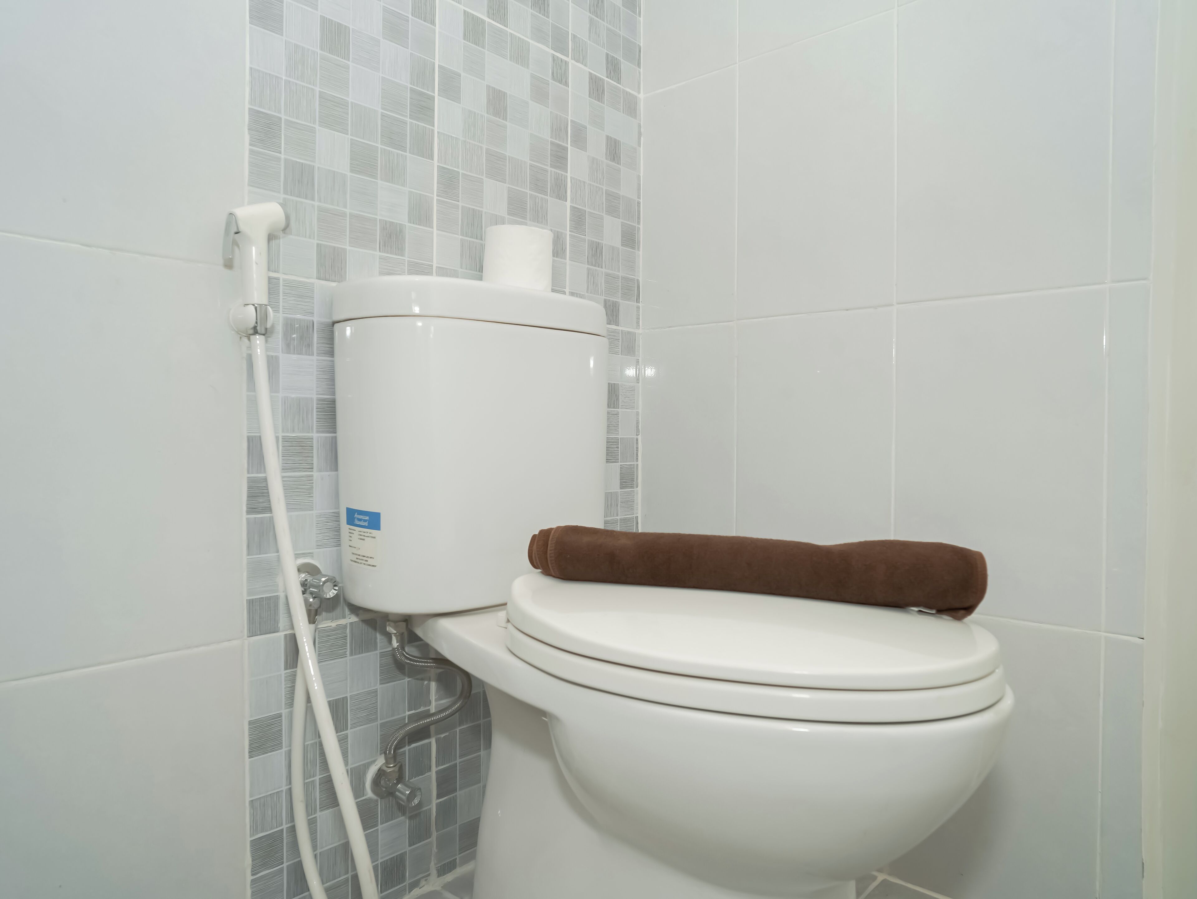 Des toilettes avec une serviette marron posée sur le siège, un porte-rouleau de papier toilette blanc et un mur carrelé à motifs à carreaux.
