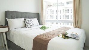 Uma cama bem arrumada com cabeceira cinza, roupa de cama branca e um cobertor marrom. Há uma mesa de cabeceira com um controle remoto e uma pequena planta.