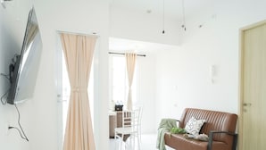 Uma sala de estar moderna com uma TV de tela plana na parede, um sofá de couro marrom com almofada, uma mesa de jantar com cadeiras e uma porta de correr com cortinas bege.