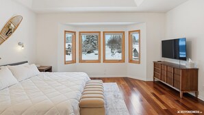 A bedroom with a large bed, a TV, a wooden cabinet, and a view of a snowy landscape through four windows. - 2BR 2BA Quiet Coastal Trail Retreat Near Downtown & Medical Centers (Anchorage)