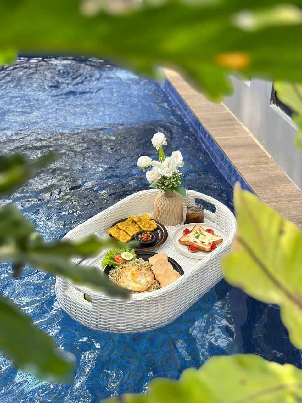 A poolside setting with a wicker basket containing food and drinks, surrounded by greenery.
