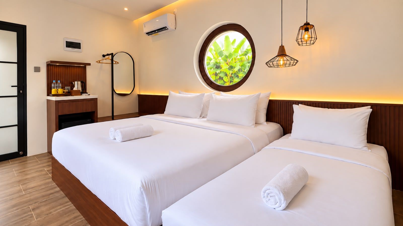 A hotel room with two beds, a small desk, a mirror, an air conditioning unit, and a window with a view of greenery.