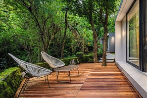 A wooden deck with two white chairs, surrounded by lush greenery and trees. - Guru Giri Forest Cabin Villa Yogyakarta (Srimulyo)