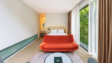 A modern hotel room with a bed, a sofa, a round table, and a view of greenery through large windows.