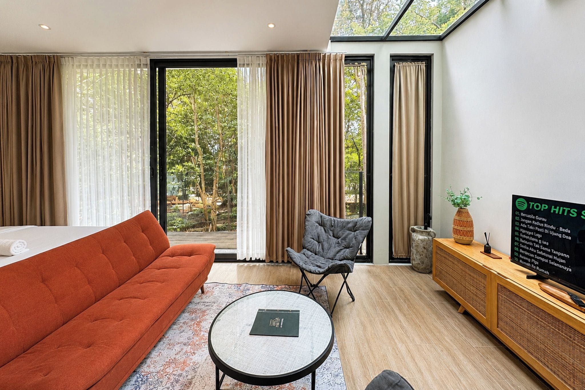 A modern hotel room with a sofa, a small table, a TV, and a view of greenery through large windows.