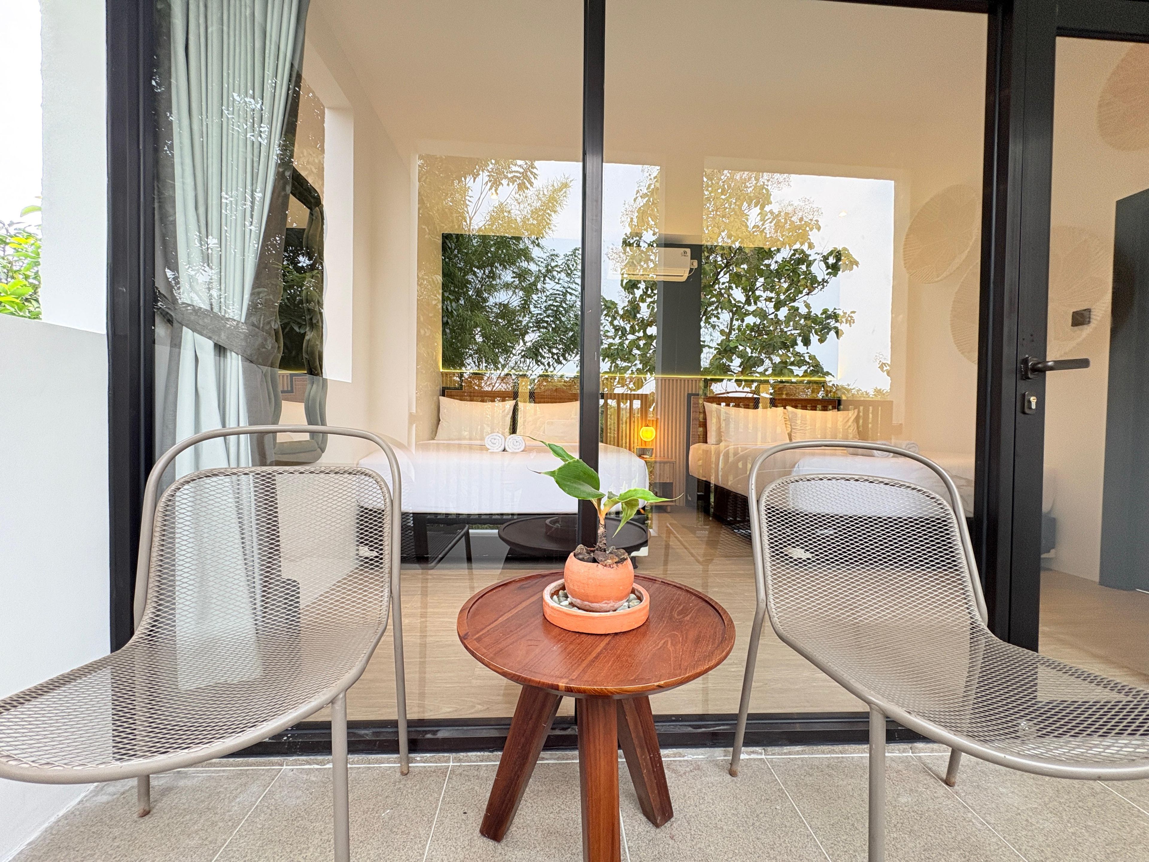 Una habitación con cama, dos sillas, una mesita con una planta y vista a un jardín a través de una puerta de vidrio.