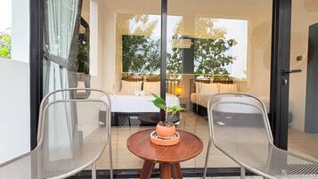 Una habitación con cama, dos sillas, una mesita con una planta y vista a un jardín a través de una puerta de vidrio.