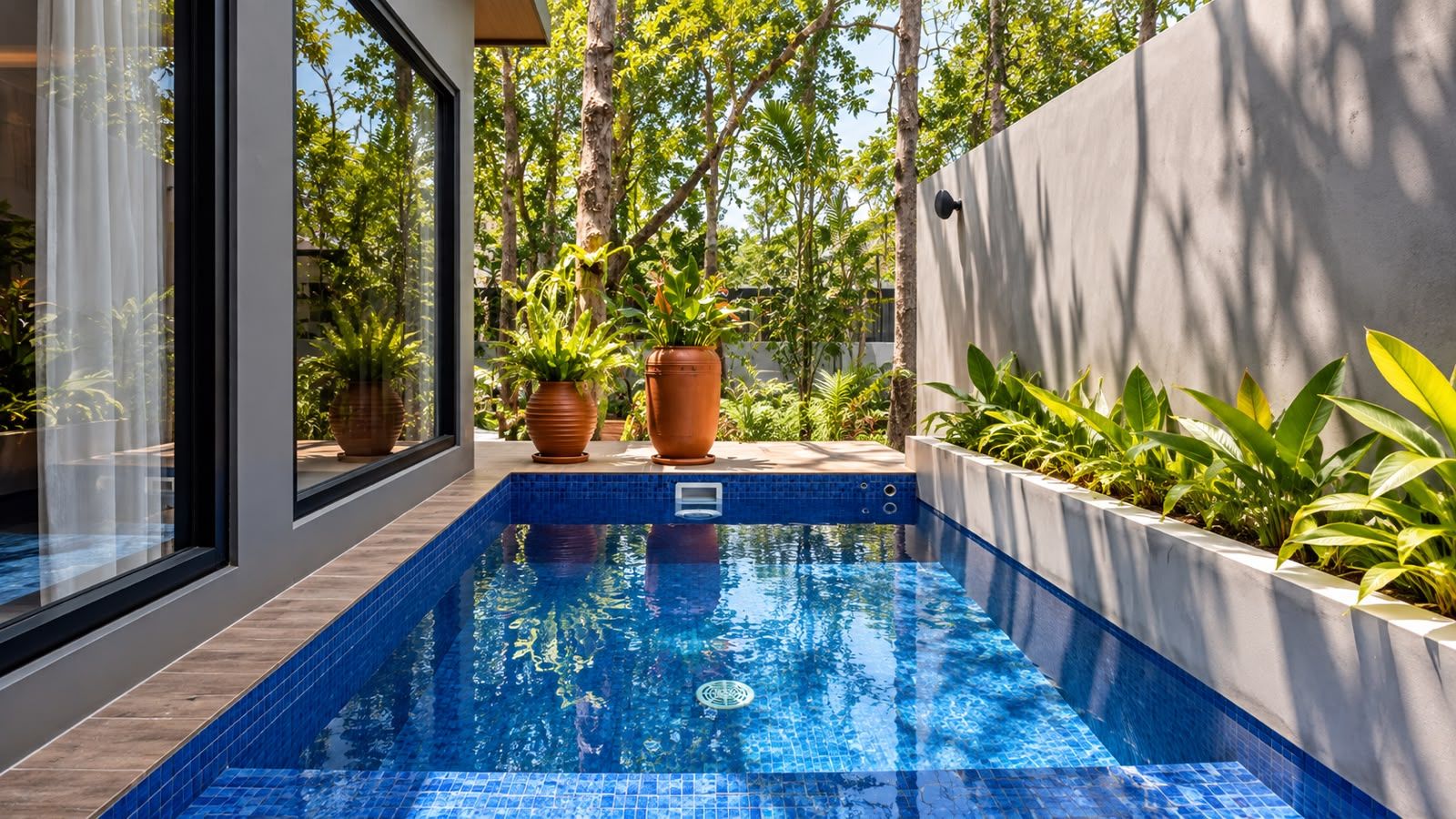 A rectangular swimming pool with blue tiles, surrounded by a wooden deck and potted plants.