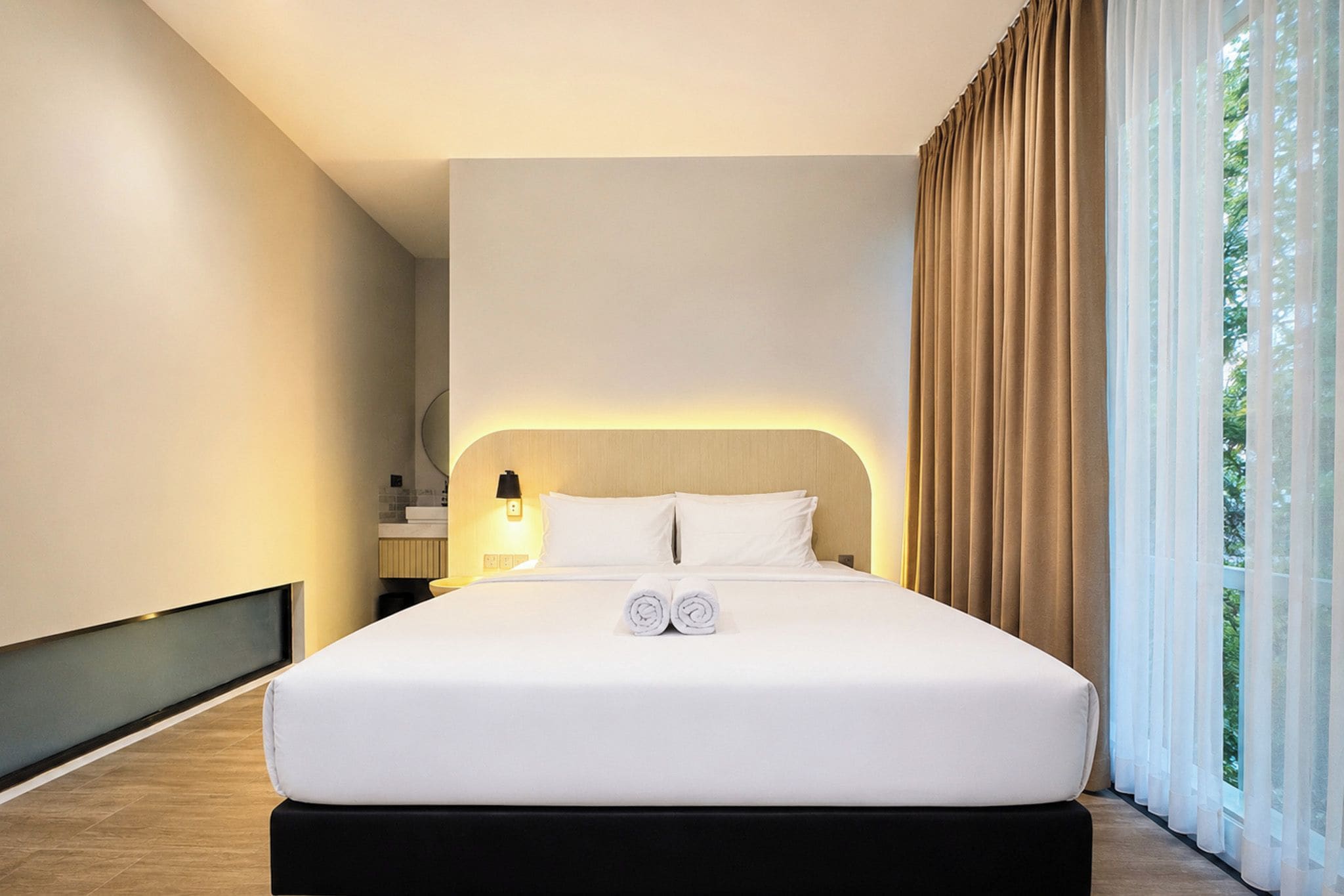 Een moderne hotelkamer met een groot bed, een bedlampje en uitzicht op groen door dunne gordijnen.