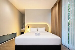 Ein modernes Hotelzimmer mit einem großen Bett, einer Nachttischlampe und Blick auf Grün hinter transparenten Vorhängen.