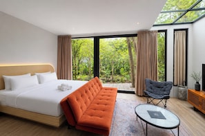 Habitación de hotel moderna con una cama grande, un sofá, una silla, una mesita y vistas a zonas verdes a través de amplios ventanales.