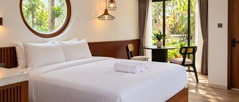 A hotel room with a large bed, a round window with a view of greenery, a small table with a chair, and a hanging light fixture.